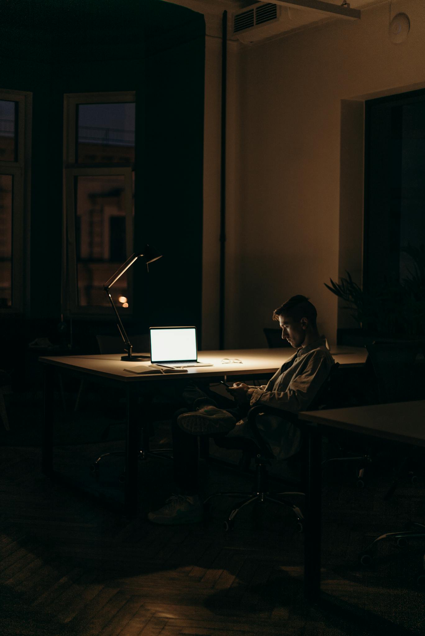 A lone man working on a laptop under dim lighting in a quiet office at night.