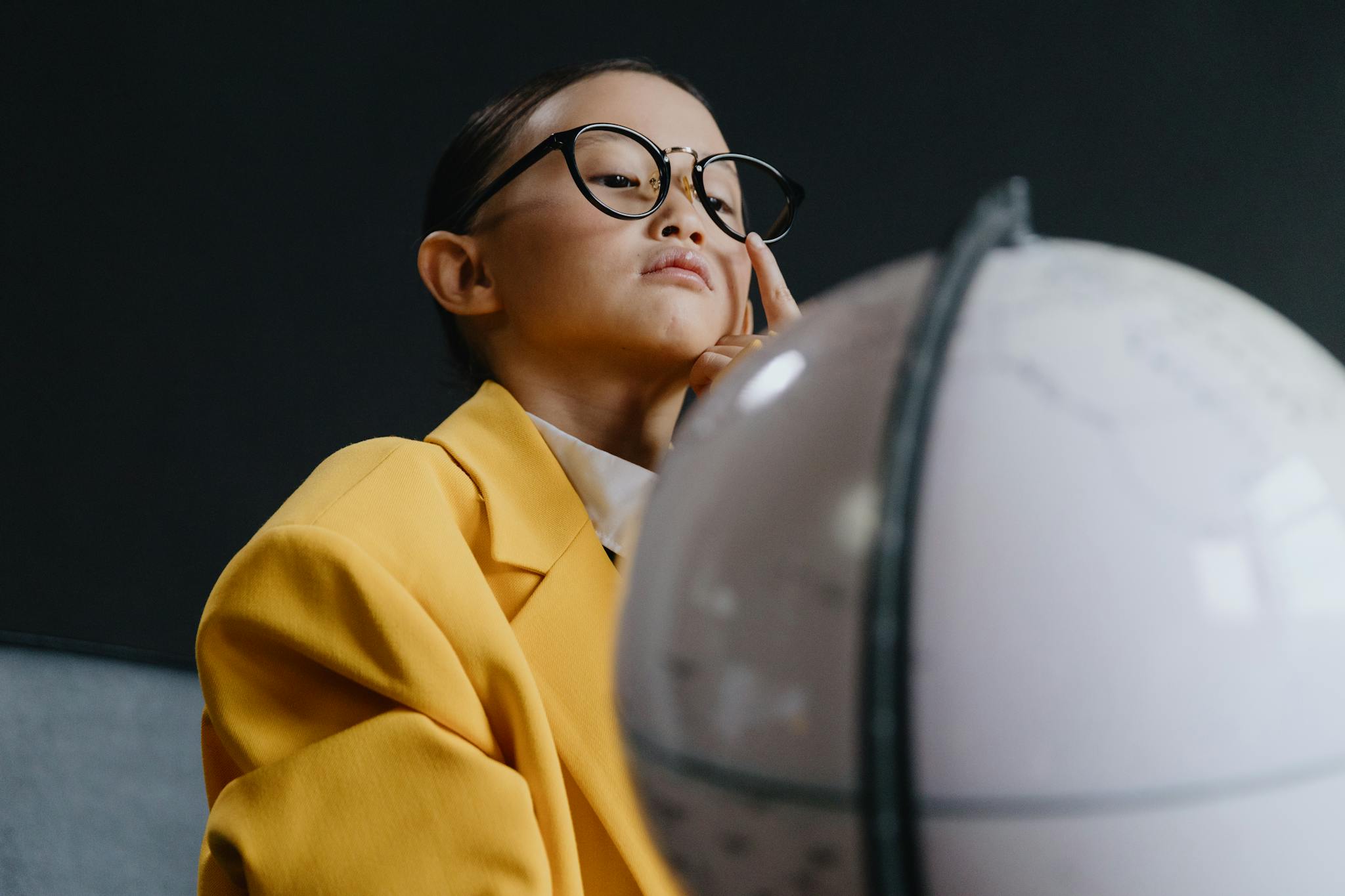 A thoughtful child in a yellow coat examining a globe indoors.