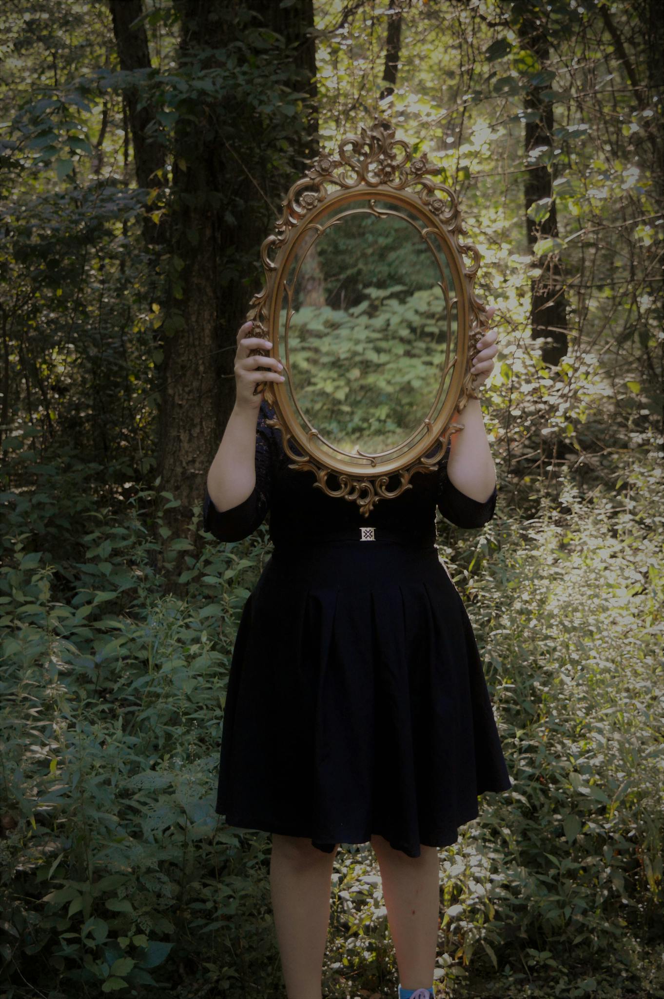 A woman holds an ornate mirror in a forest setting, reflecting nature.