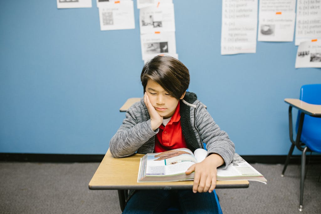 A young boy deep in thought while reading a book alone in a classroom.