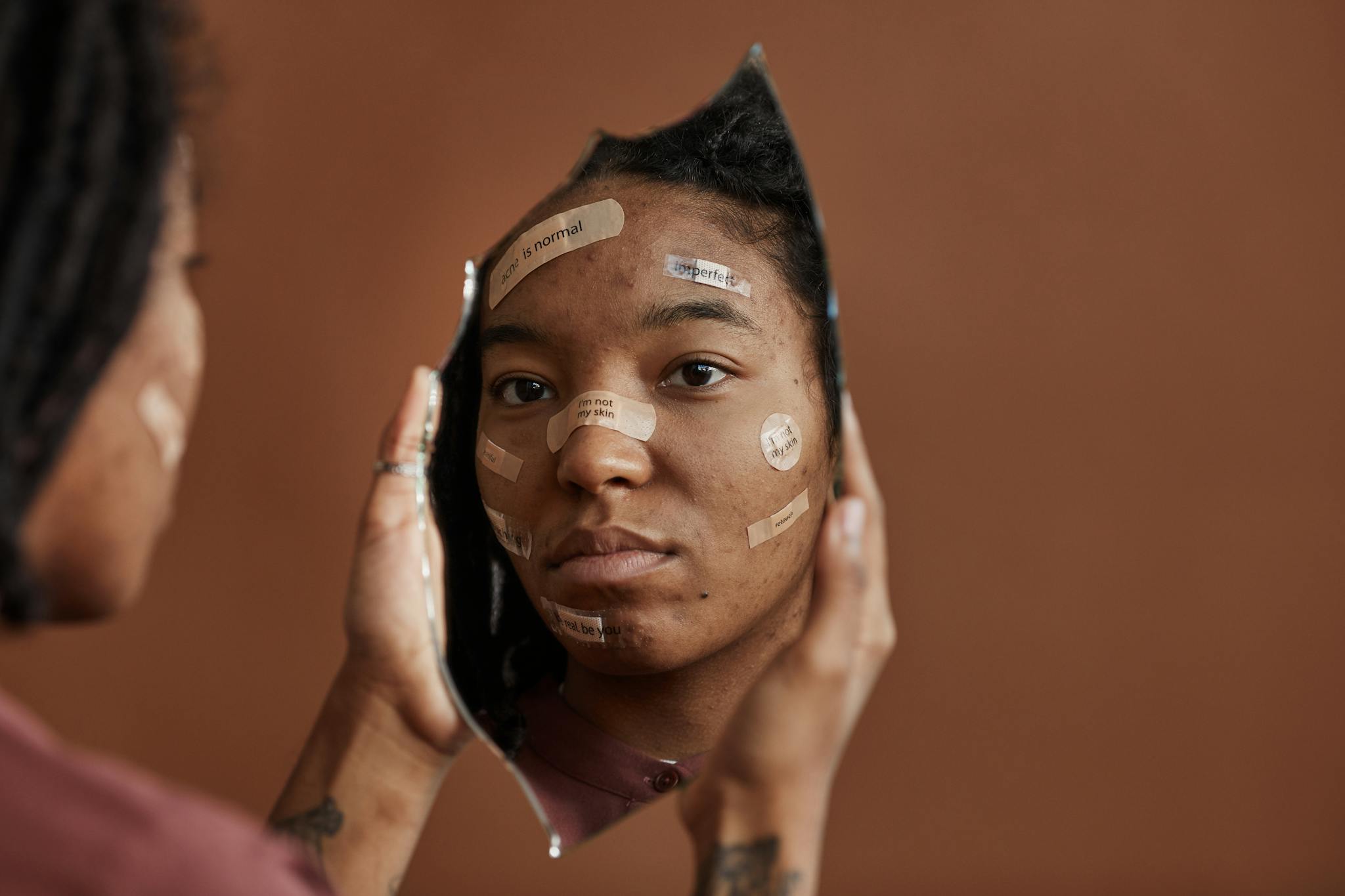 African American woman with Band-Aids looks into a shattered mirror, reflecting self-acceptance.