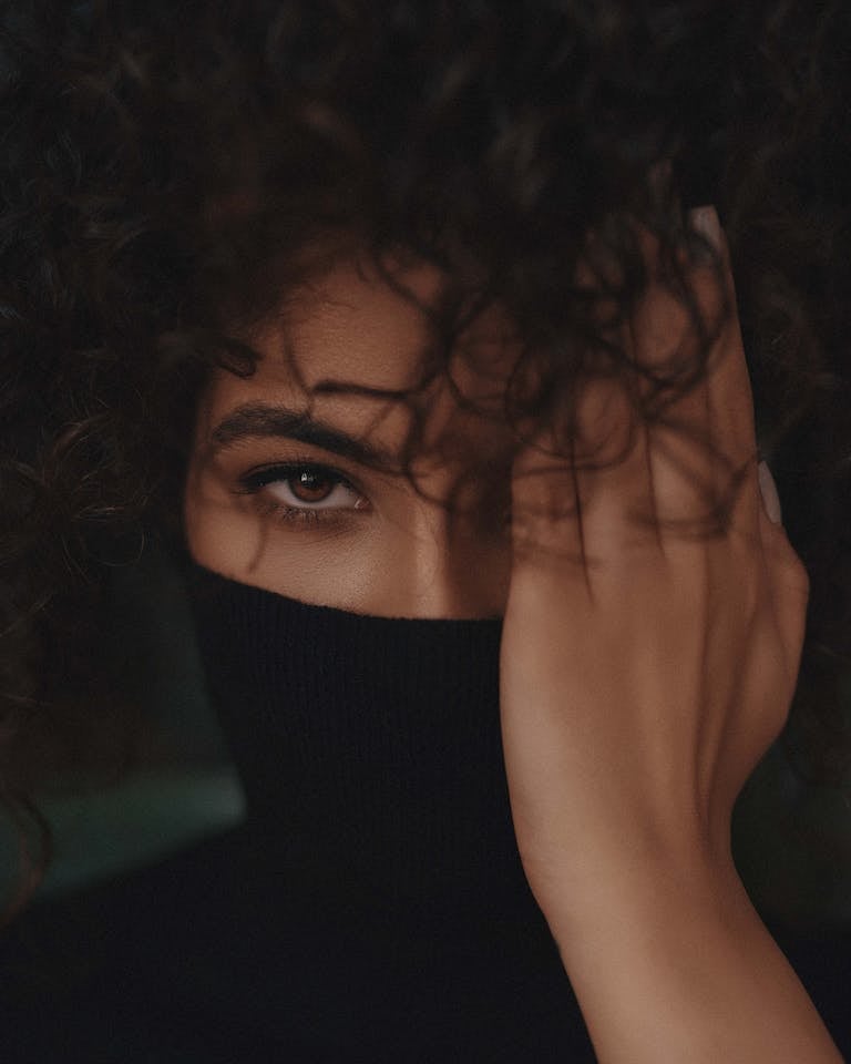 Close-up of a woman with curly hair, partially hiding her face with a black turtleneck and hand.