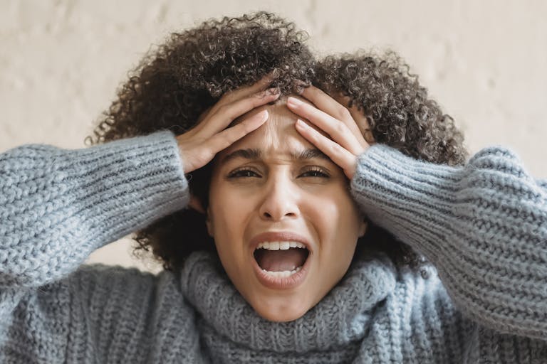 Expressive African American woman with curly brown hair touching head and yelling against light background