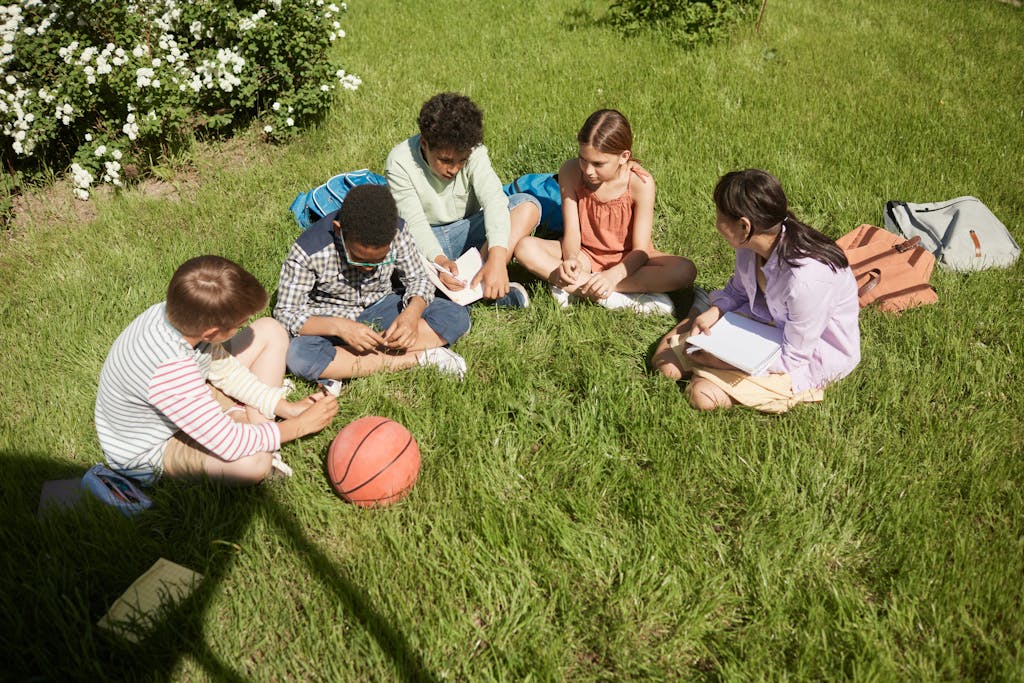 Five teenagers sitting on grass, studying together in a park on a sunny summer day.