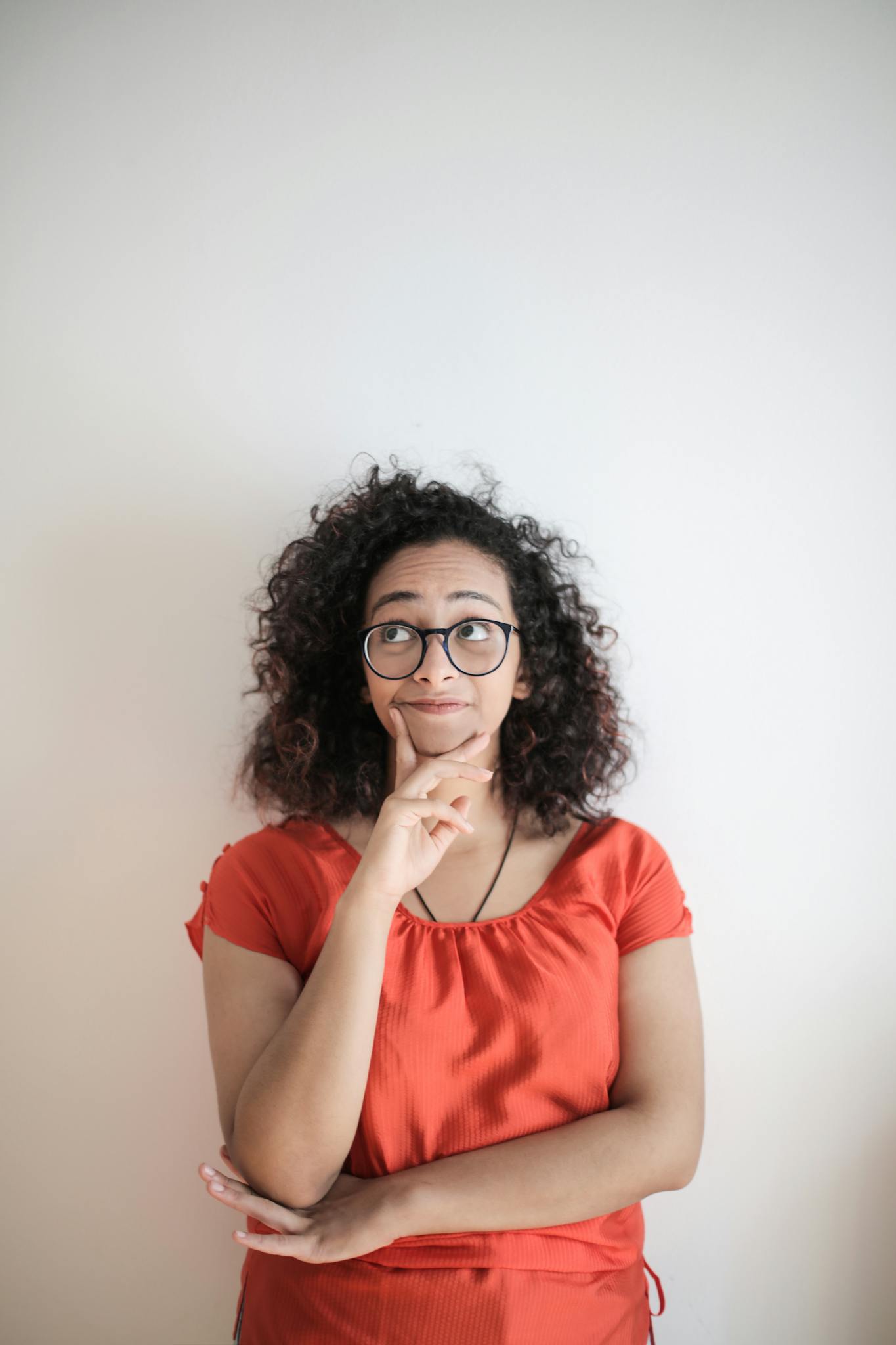 Portrait of a thoughtful woman in a red top, with curly hair and glasses against a white background.