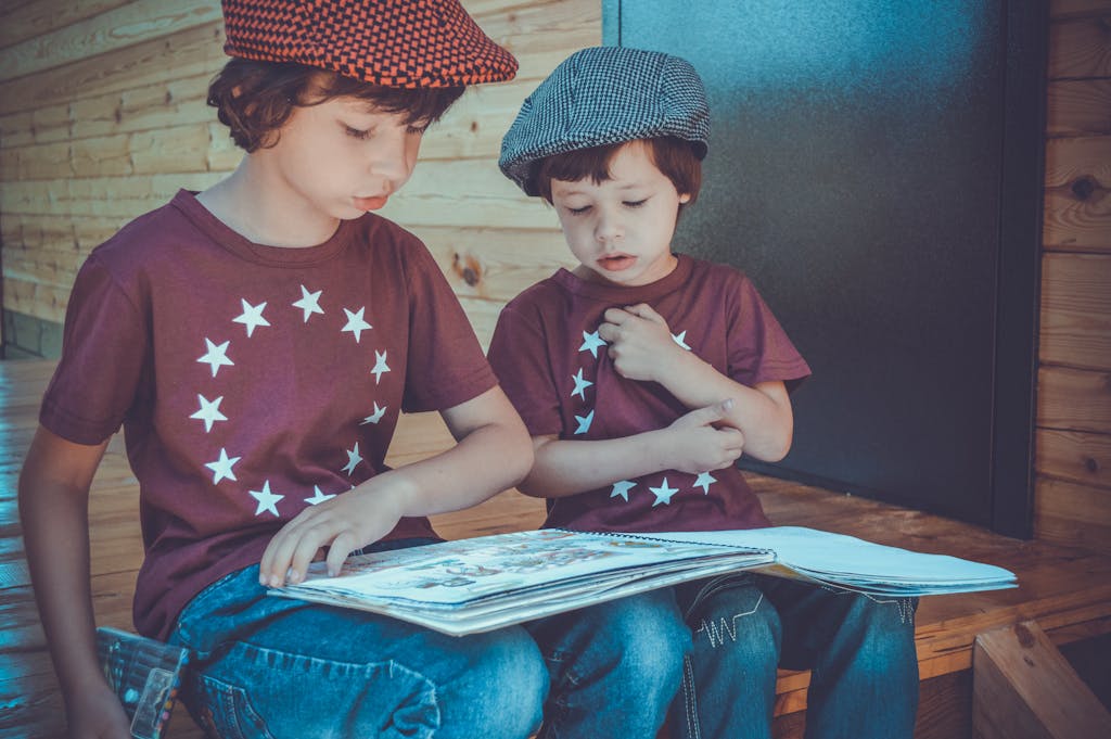 Two young boys wearing hats and star shirts reading a book outdoors on a wooden porch.